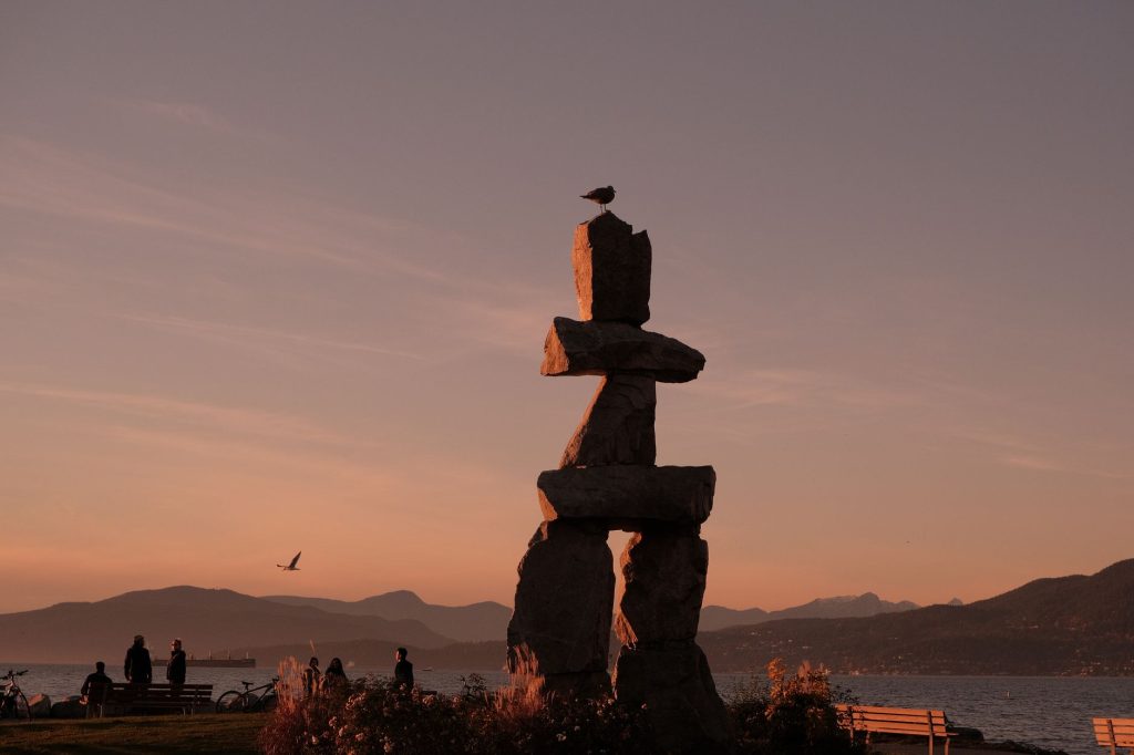 Silhouette d'un inukshuk se détachant sur un ciel rose de coucher de soleil, au-dessus d'un paysage côtier.