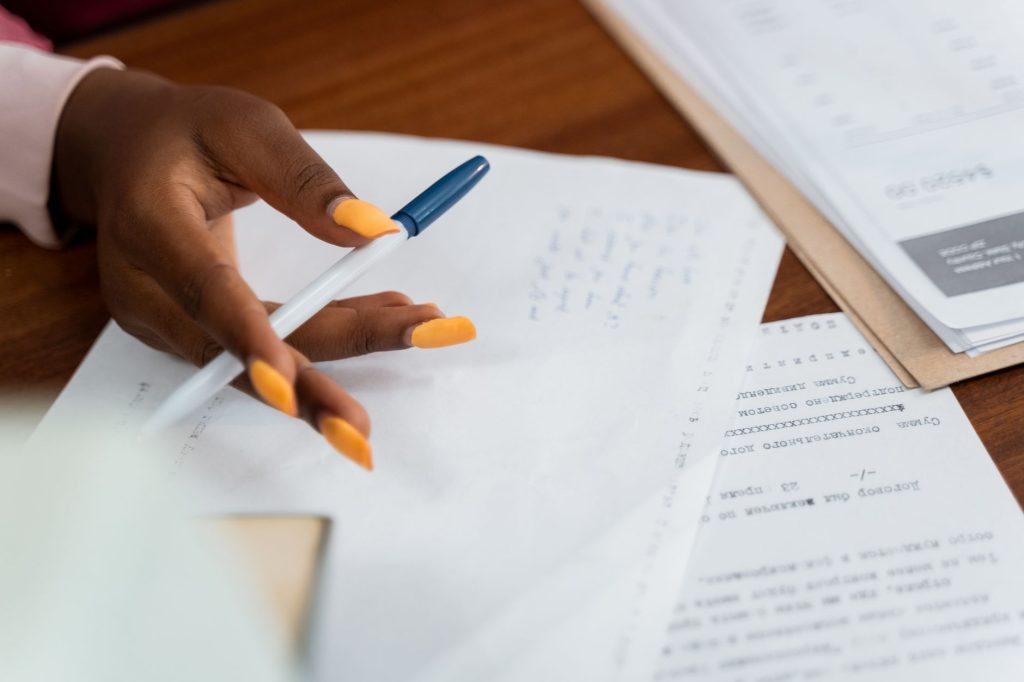 Une main sur des documents sur un bureau, avec un stylo et des notes écrites, évoque un environnement de travail occupé.